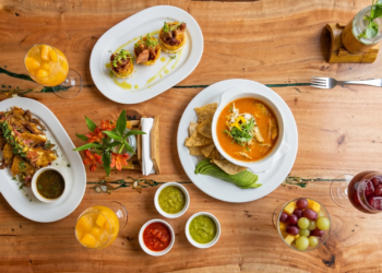 Una mesa con platos de cocina fusión latina: sopa de tortilla, tacos, patacones rellenos y bebidas frutales sobre madera rústica.