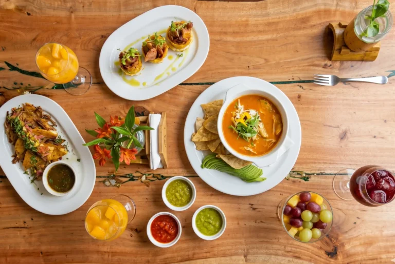 Una mesa con platos de cocina fusión latina: sopa de tortilla, tacos, patacones rellenos y bebidas frutales sobre madera rústica.