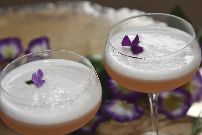 Dos copas de Pisco Sour con espuma cremosa y flores púrpuras comestibles, servidos en la barra del 3 Soles Restaurant.