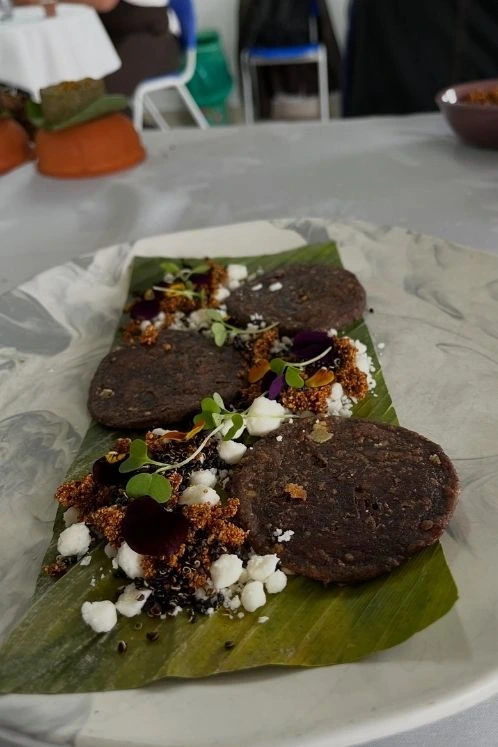 Tres arepas artesanales de maíz morado servidas sobre una hoja de plátano con queso fresco, quinua crujiente y brotes verdes.